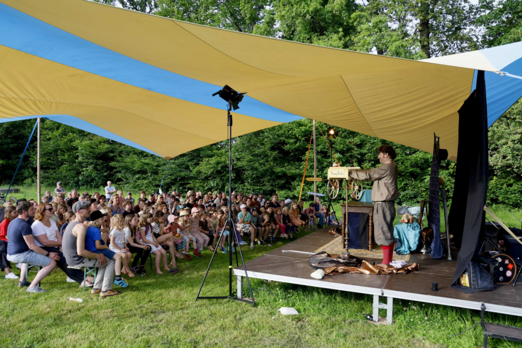 Ein Mensch auf einer Bühne, vor der eine Gruppe Menschen, vor allem Kinder, sitzt. Über der Bühne ist ein buntes Tarp gespannt, im Hintergrund Wald und Wiese.