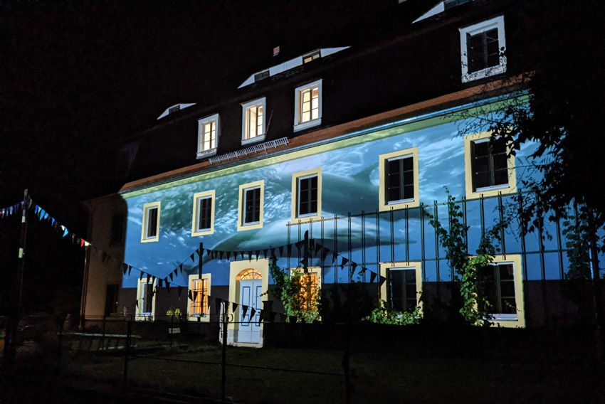 Ein Haus im Dunkeln, dessen Fassade durch eine Licht-Projektion beleuchtet ist. Die Projektion zeigt Delfine unter Wasser.