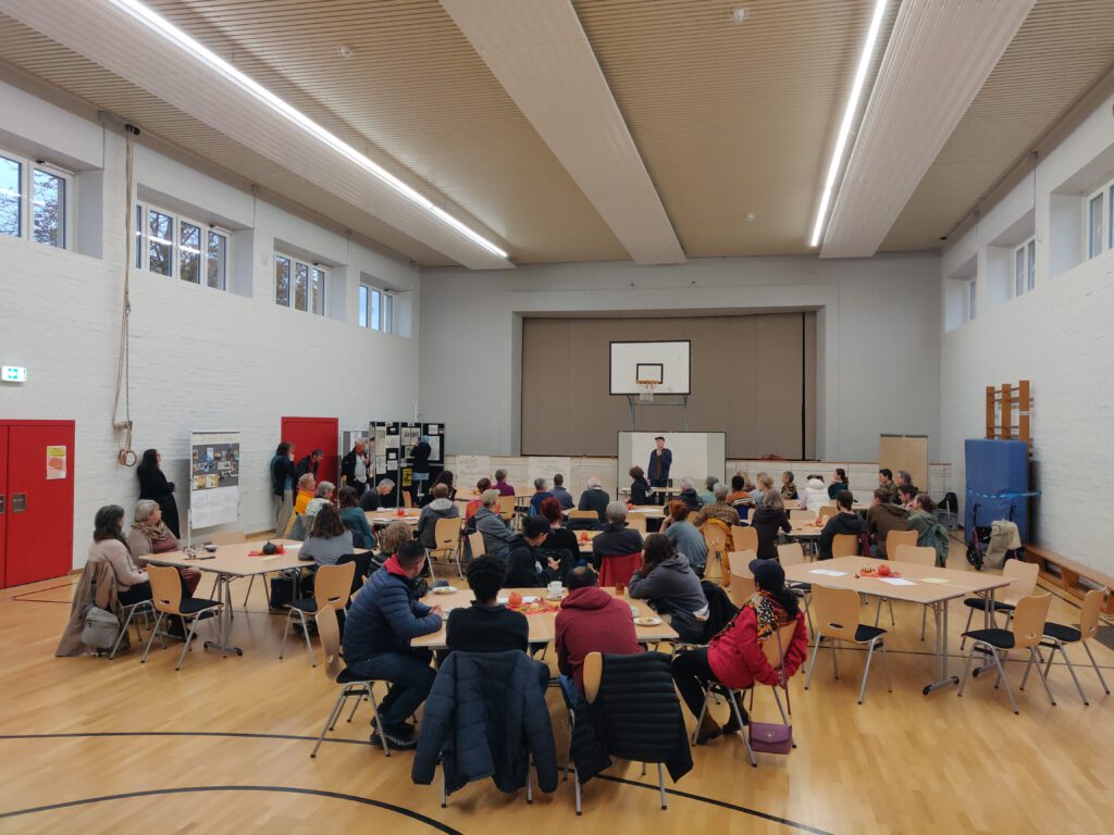 Eine Turnhalle, in der Menschen sich versammelt haben. Die meisten sitzen in Tischgruppen, einige stehen am Rand neben einer aufgestellten Pinnwand, eine Person steht vorne.