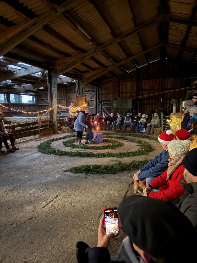 Blick in einen geräumigen Stall, in dem eine Spirale aus Tannenzweigen liegt, in der Mitte steht eine Kerze. Drum herum sitzen Menschen auf Strohballen und der Raum ist mit Lichterketten geschmückt.