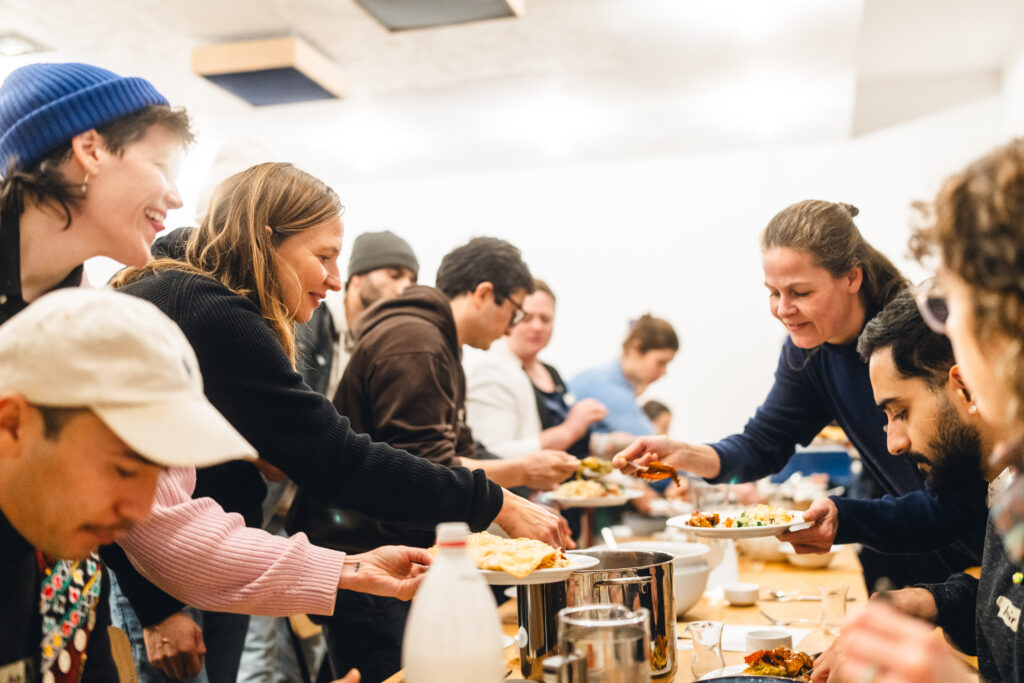 Mehrere Personen stehen und sitzen an einem langen Tisch und teilen sich gerade Essen auf Teller aus. Auf dem Tisch befindet sich ein großer Topf, Gläser und Besteck.
